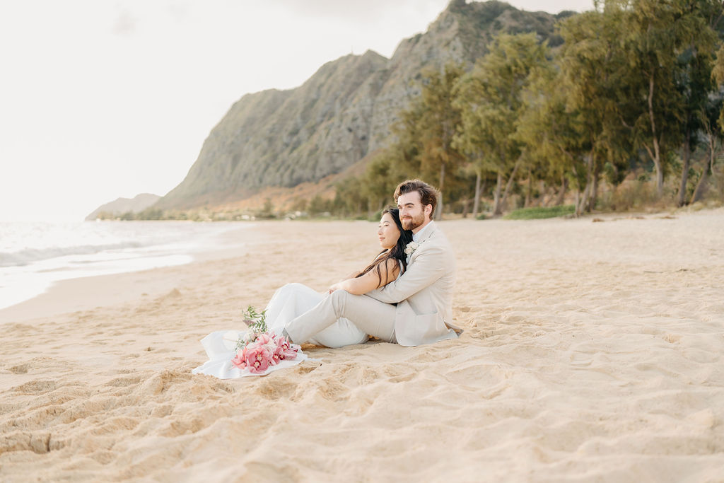 A couple in wedding attire embraces with the ocean and sky in the background. The woman holds a bouquet