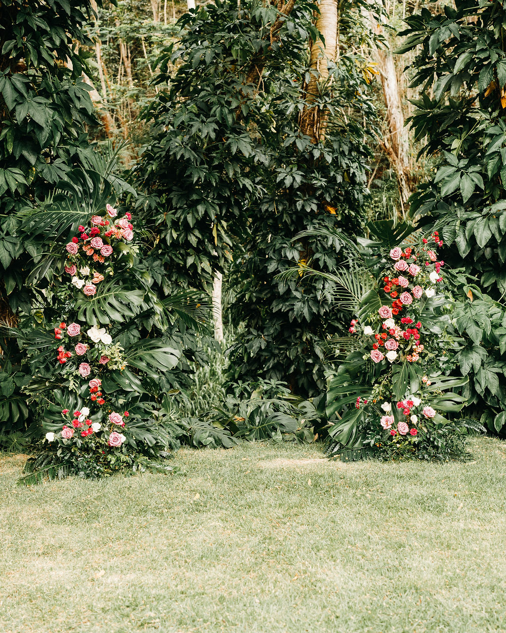 Two vertical floral arrangements with pink and red flowers and greenery are set outdoors in front of dense green foliage.
