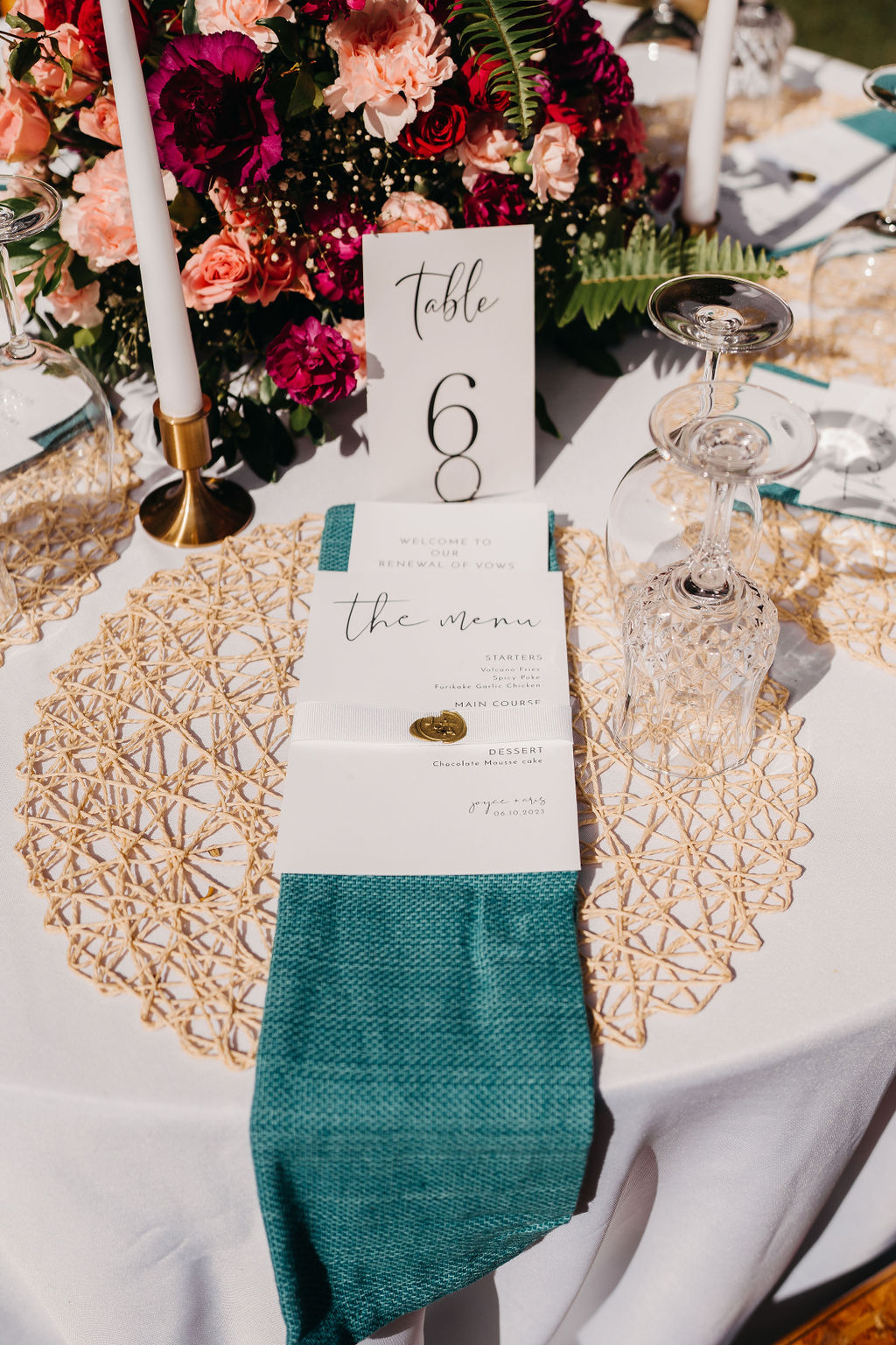 A wedding table setup with a gold placemat, teal napkin, menu card, table number sign reading "Table 6," wine glasses, and floral centerpiece with pink and red flowers.