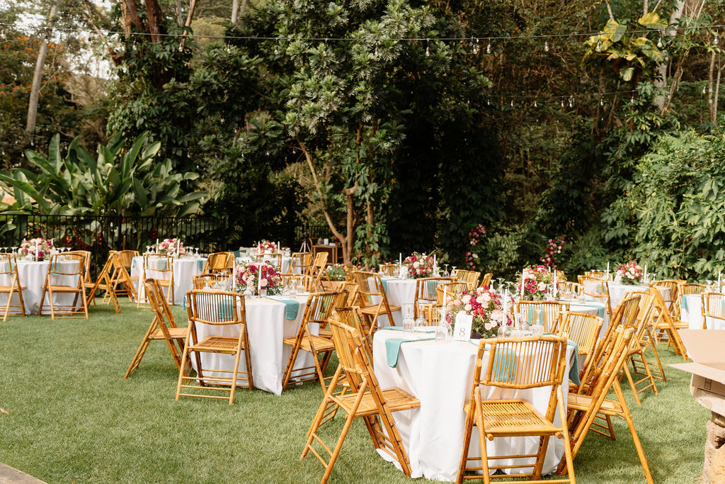 Round tables with white tablecloths and floral centerpieces are arranged on a grassy lawn, surrounded by wooden chairs and greenery.