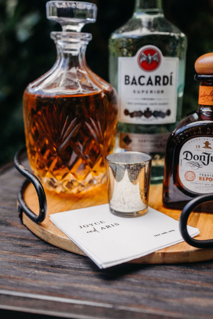 A wooden tray holding a crystal decanter with brown liquor, bottles of Bacardi rum and Don Julio, a metal jigger, and a notepad reading “LOVE & MAKERS” on a table.