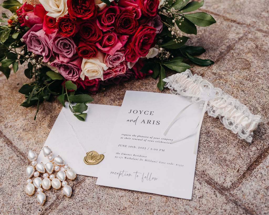 A wedding invitation, pearl jewelry, white lace garter, and a bouquet of red, pink, and white roses are arranged on a stone surface.
