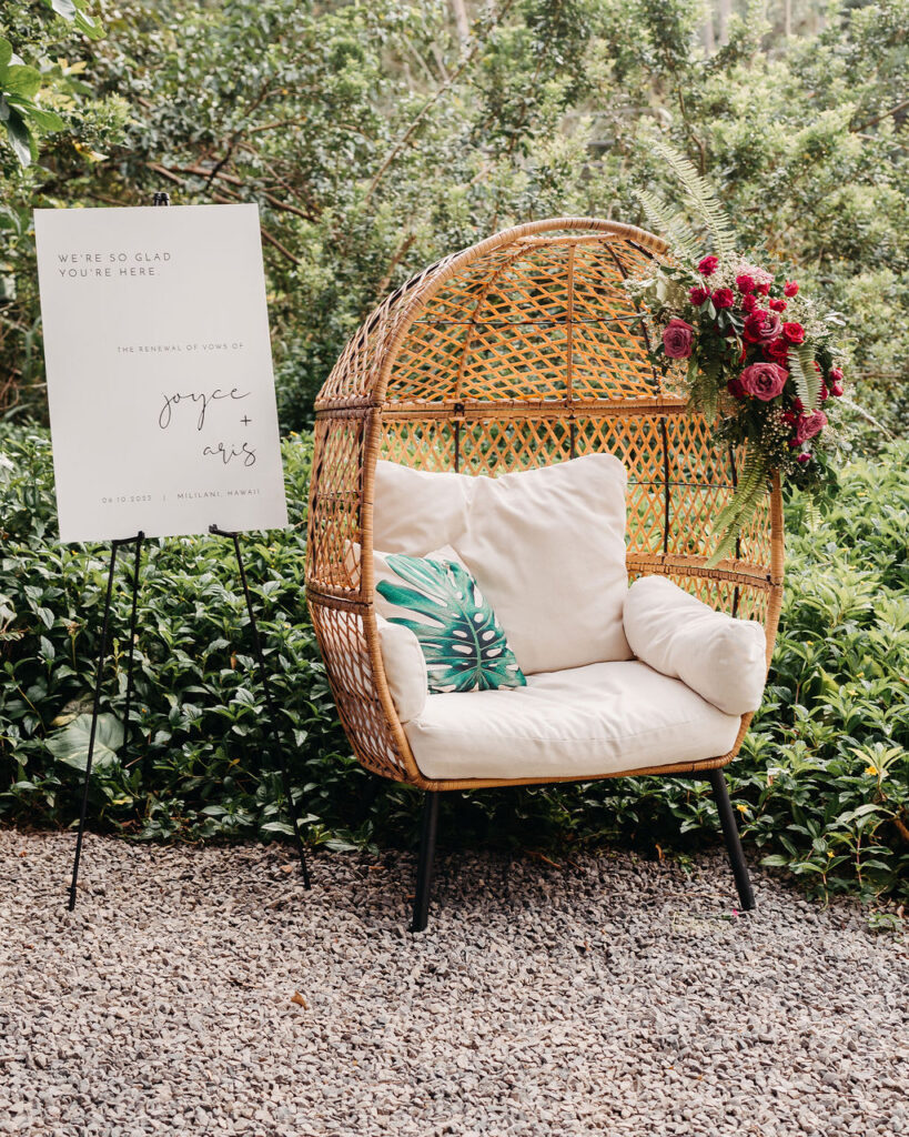 A wicker chair with white cushions and a green leaf pillow is set on gravel next to an easel with a white sign; a bouquet of flowers decorates the chair. Shrubs and trees are in the background.