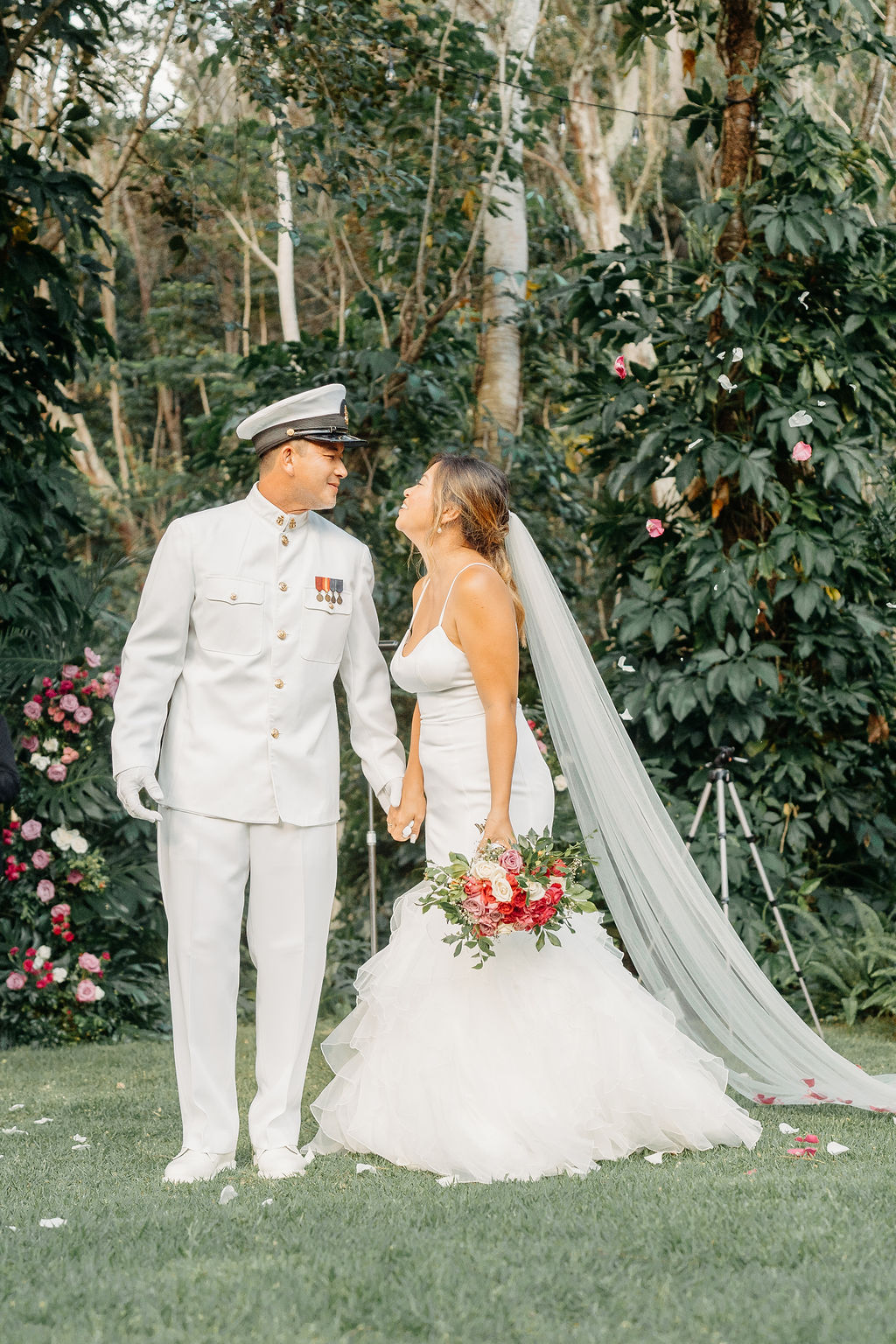 A bride in a white dress and a groom in a white military uniform stand together on a forested path, smiling and holding a bouquet for their backyard wedding in Oahu