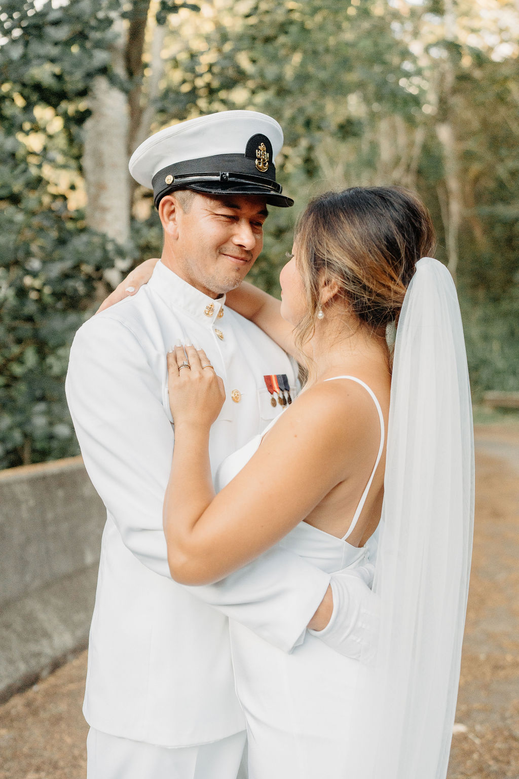A bride in a white dress and a groom in a white military uniform stand together on a forested path, smiling and holding a bouquet for their backyard wedding in Oahu