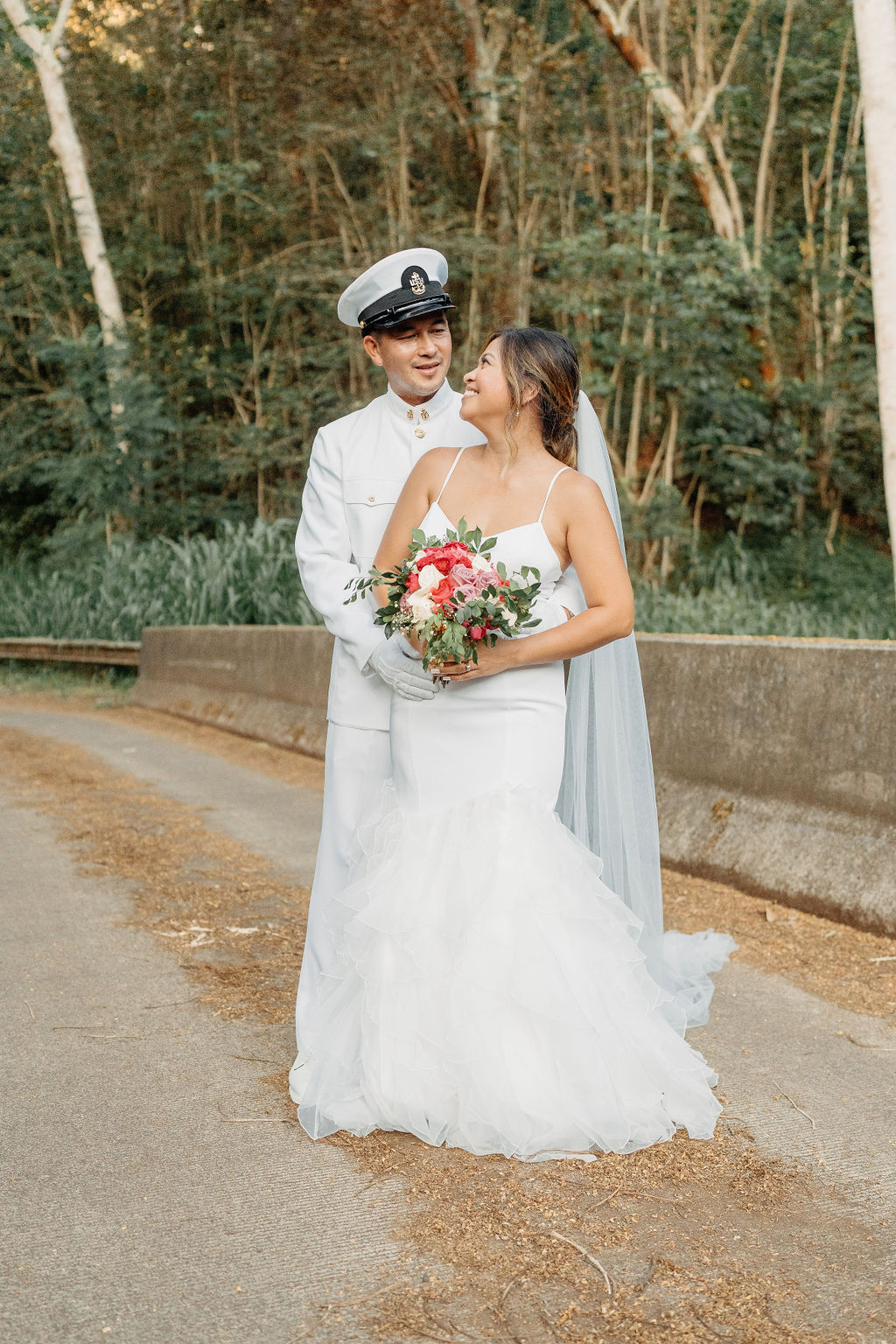 A bride in a white dress and a groom in a white military uniform stand together on a forested path, smiling and holding a bouquet for their backyard wedding in Oahu