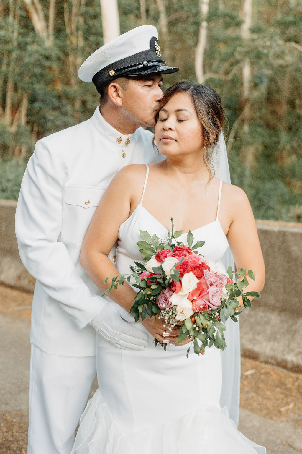A bride in a white dress and a groom in a white military uniform stand together on a forested path, smiling and holding a bouquet for their backyard wedding in Oahu