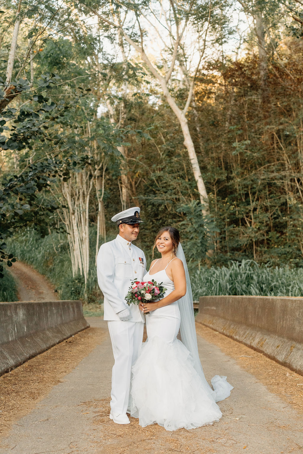 A bride in a white dress and a groom in a white military uniform stand together on a forested path, smiling and holding a bouquet for their backyard wedding in Oahu