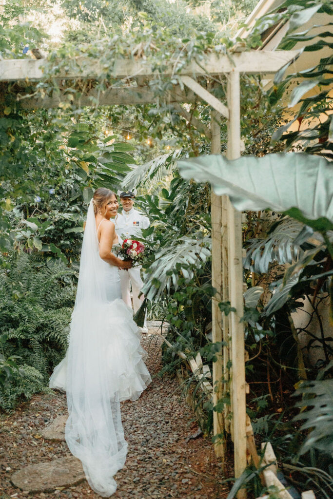 A bride in a white dress and a groom in a white military uniform stand together on a forested path, smiling and holding a bouquet for their backyard wedding in Oahu