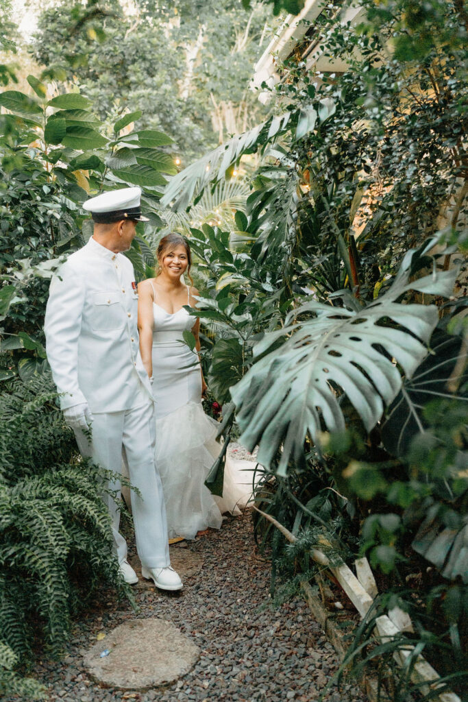 A bride in a white dress and a groom in a white military uniform stand together on a forested path, smiling and holding a bouquet for their backyard wedding in Oahu
