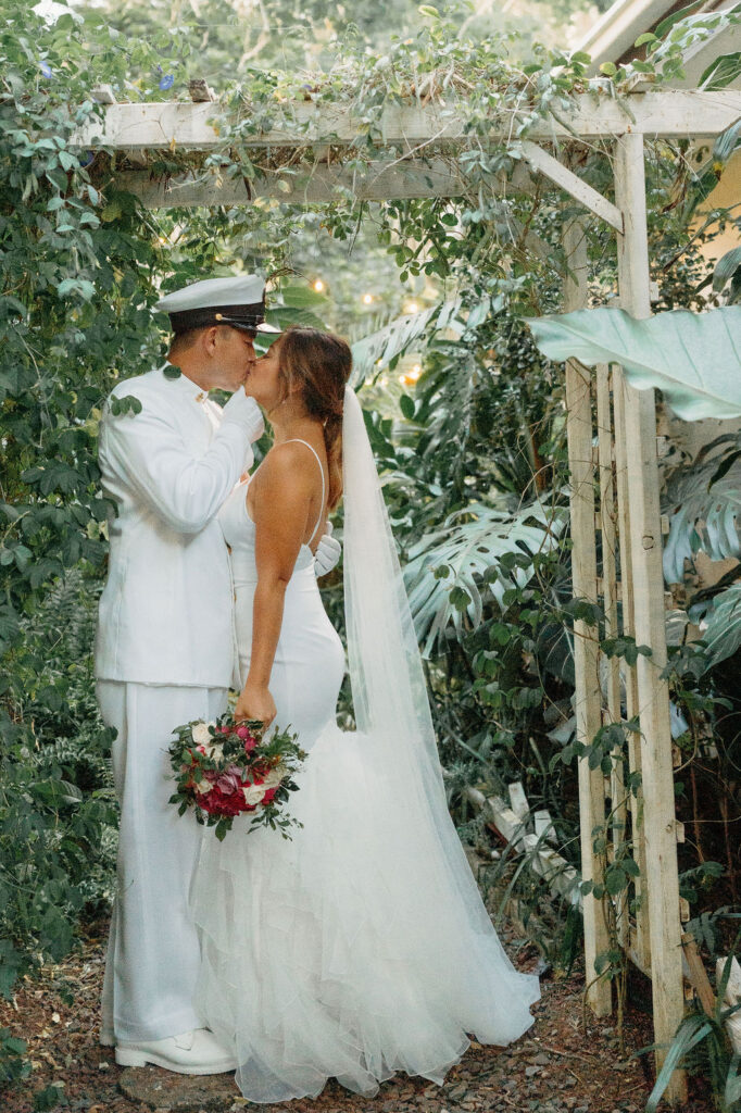 A bride in a white dress and a groom in a white military uniform stand together on a forested path, smiling and holding a bouquet for their backyard wedding in Oahu