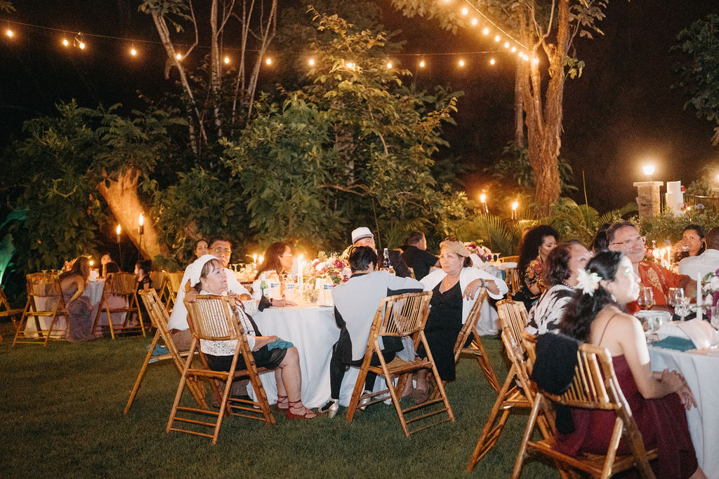 People sit around round tables at an outdoor evening event with string lights overhead, surrounded by trees and greenery for a dreamy backyard wedding in oahu