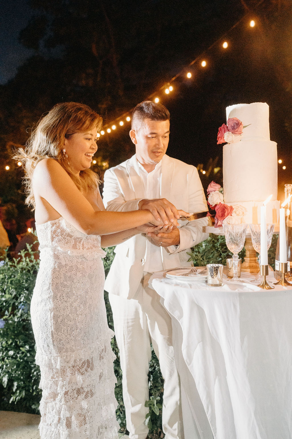 A bride and groom, both dressed in white, cut a tall white wedding cake together at an outdoor evening reception with string lights above.