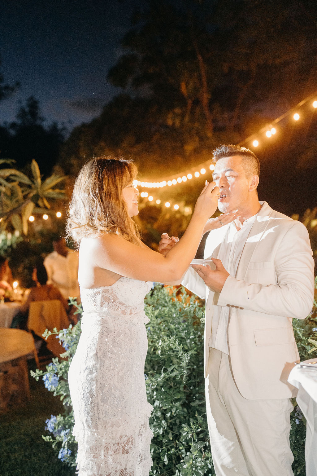 A bride and groom, both dressed in white, cut a tall white wedding cake together at an outdoor evening reception with string lights above.