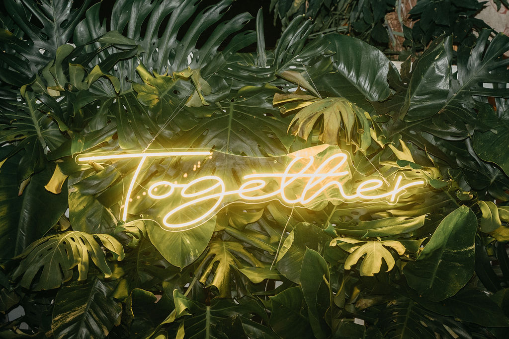 Neon sign displaying the word "Together" in cursive font, illuminated in yellow, placed against a backdrop of large green tropical leaves.