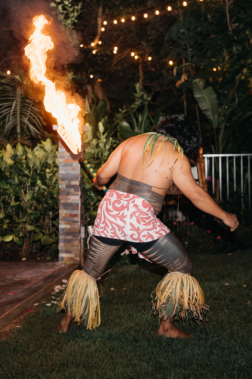 A performer in traditional attire dances with fire sticks and stands over a line of fire, while two people watch in the background outside a house at night.