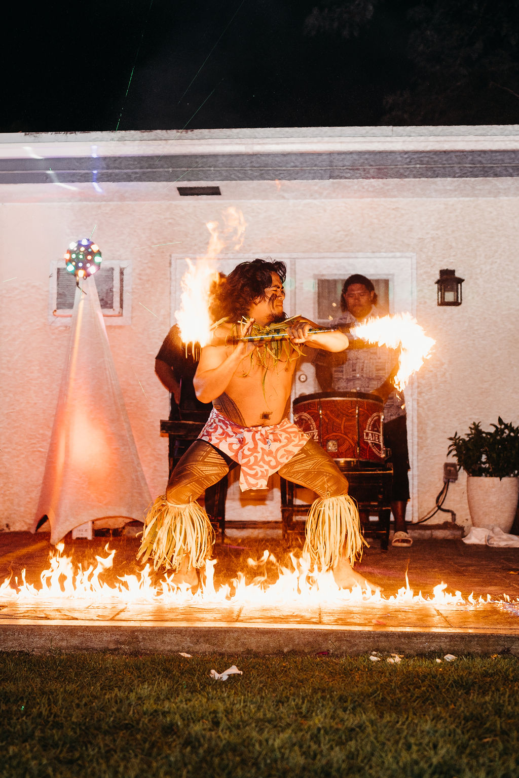 A performer in traditional attire dances with fire sticks and stands over a line of fire, while two people watch in the background outside a house at night.