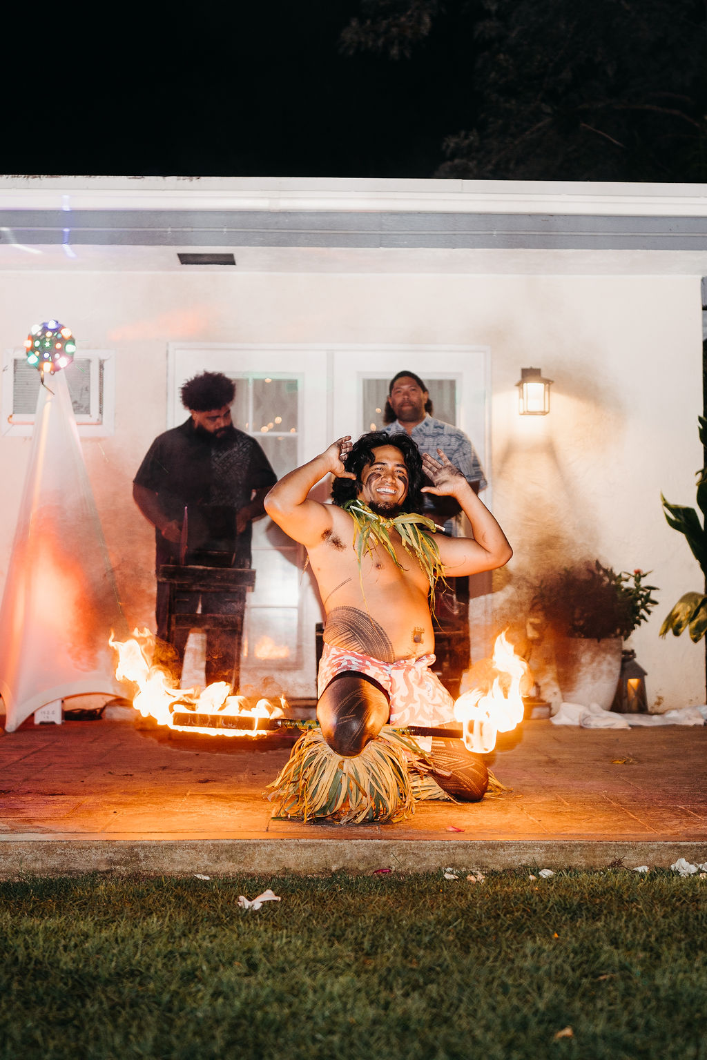 A performer in traditional attire dances with fire sticks and stands over a line of fire, while two people watch in the background outside a house at night.