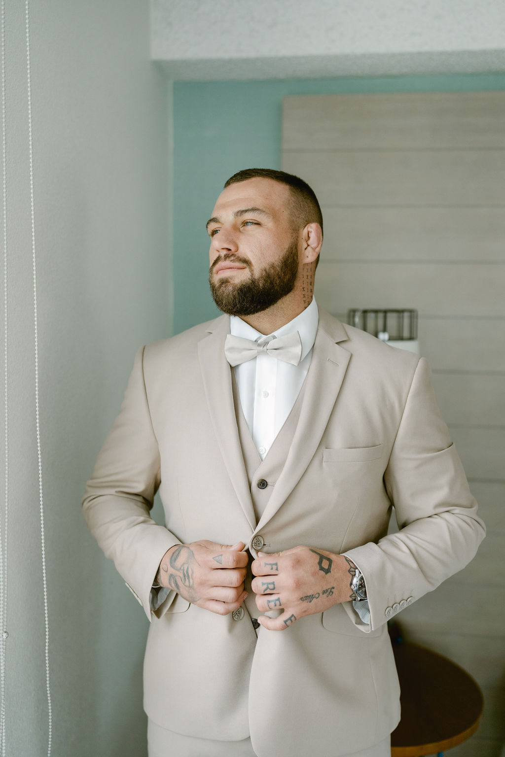 A man with tattoos adjusts his beige suit jacket and bow tie while standing indoors near a light-colored wall.