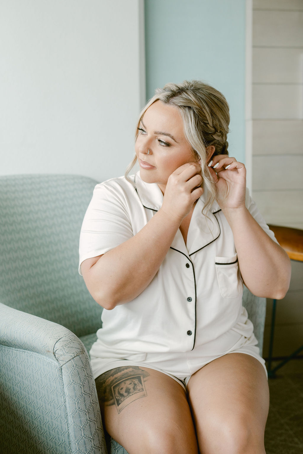 Woman sitting on a chair in light-colored pajamas, putting on an earring, with a tattoo visible on her thigh.