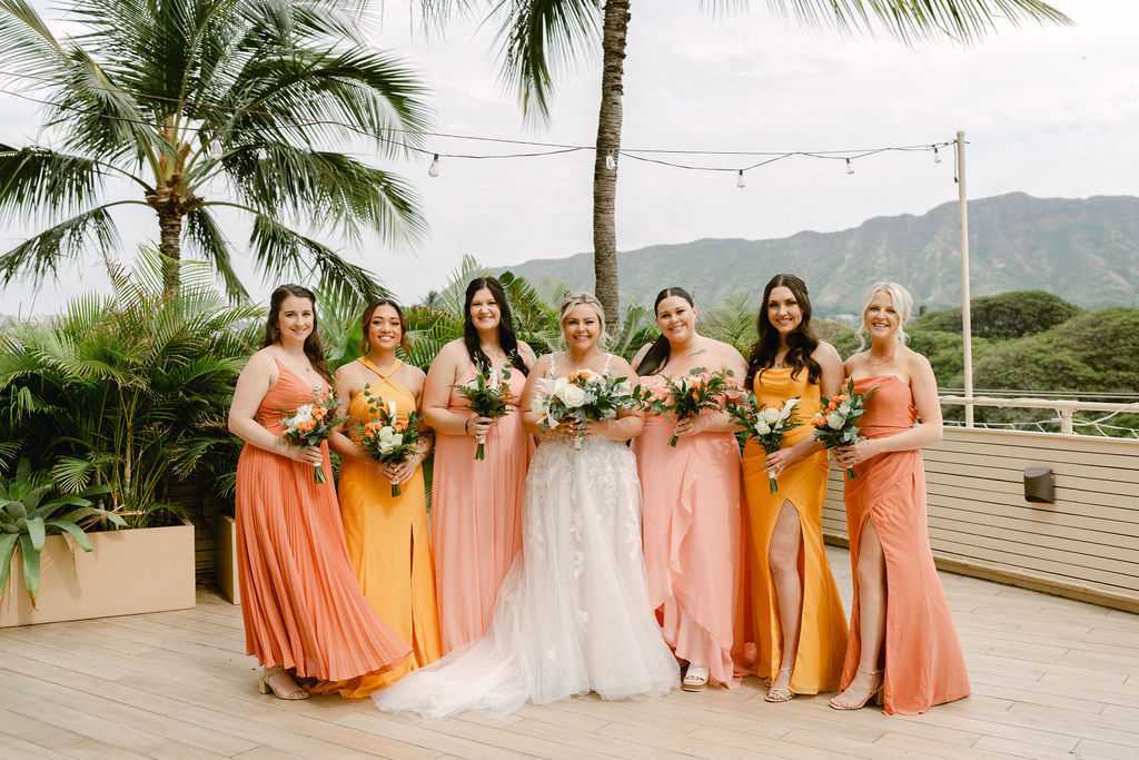 A bride and groom stand close together outdoors, surrounded by their wedding party in yellow and orange attire, with palm trees and mountains in the background for a wedding at Queen Kapiolani Hotel