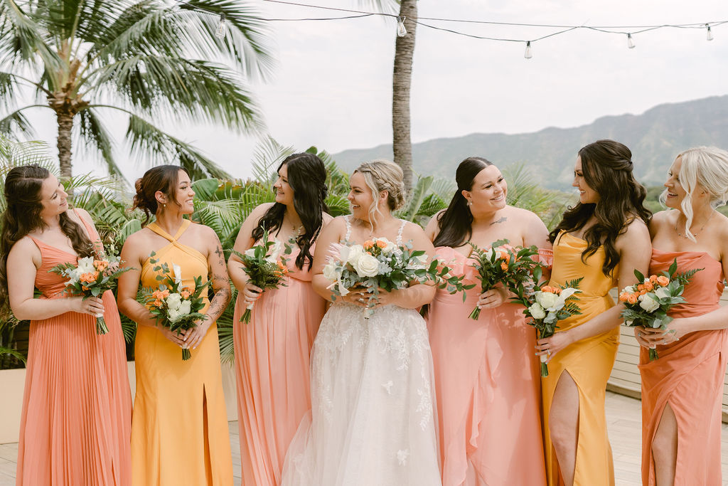 A bride and groom stand close together outdoors, surrounded by their wedding party in yellow and orange attire, with palm trees and mountains in the background for a wedding at Queen Kapiolani Hotel