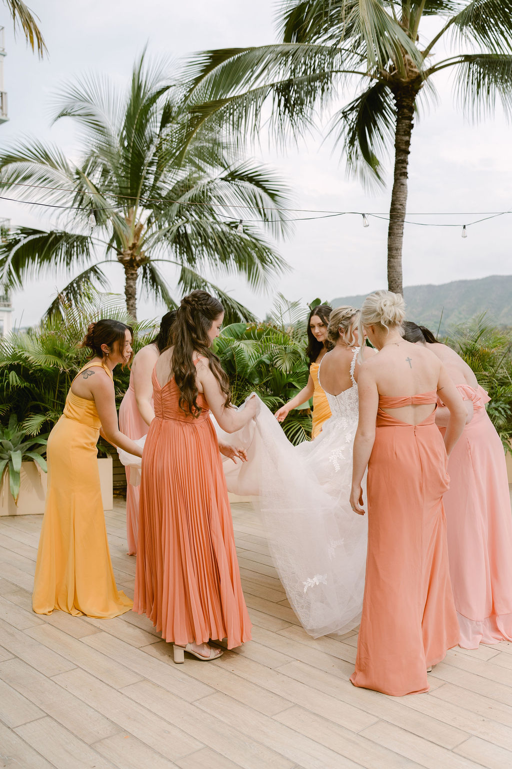 A bride and groom stand close together outdoors, surrounded by their wedding party in yellow and orange attire, with palm trees and mountains in the background for a wedding at Queen Kapiolani Hotel