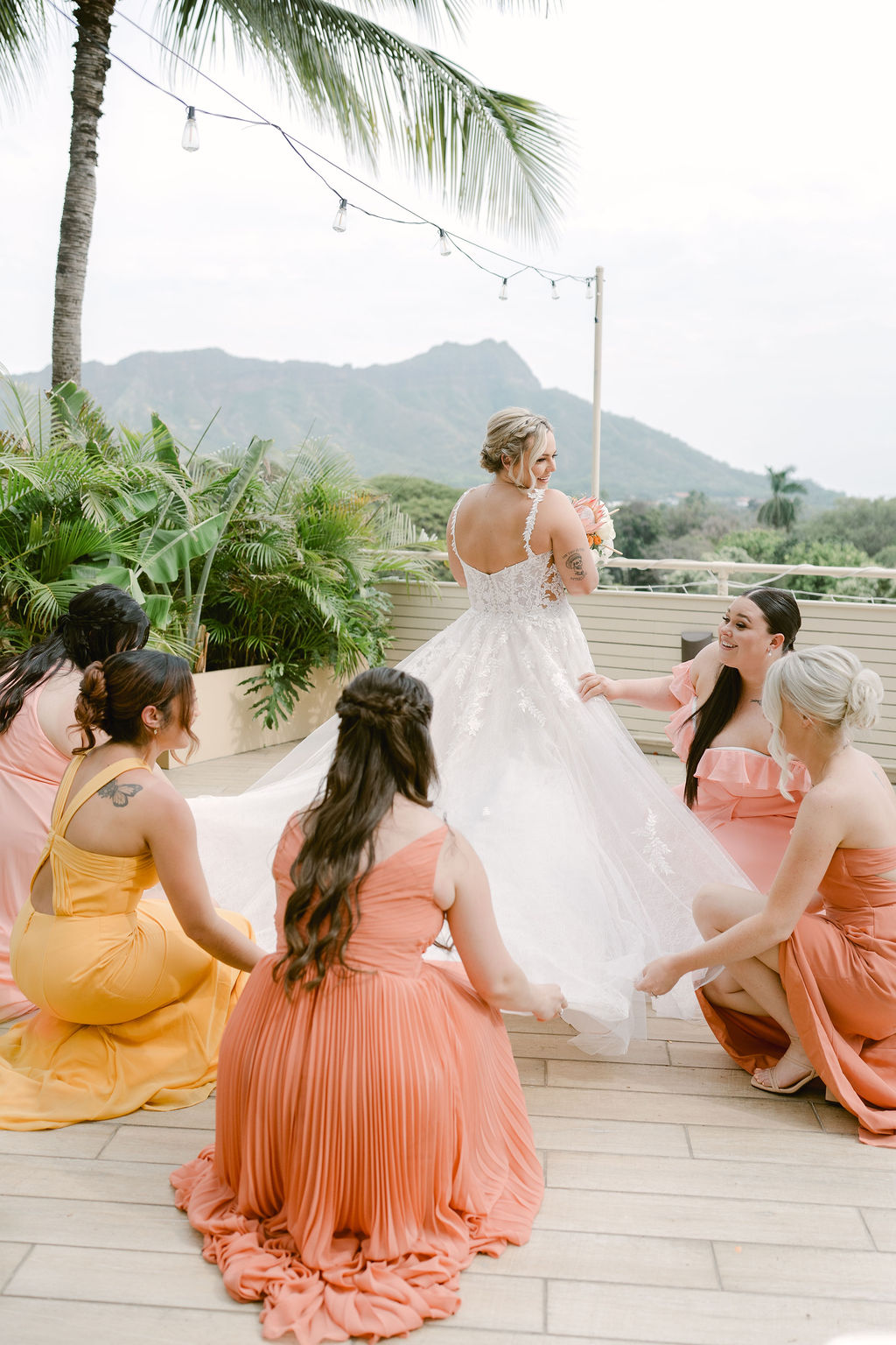 A bride and groom stand close together outdoors, surrounded by their wedding party in yellow and orange attire, with palm trees and mountains in the background for a wedding at Queen Kapiolani Hotel