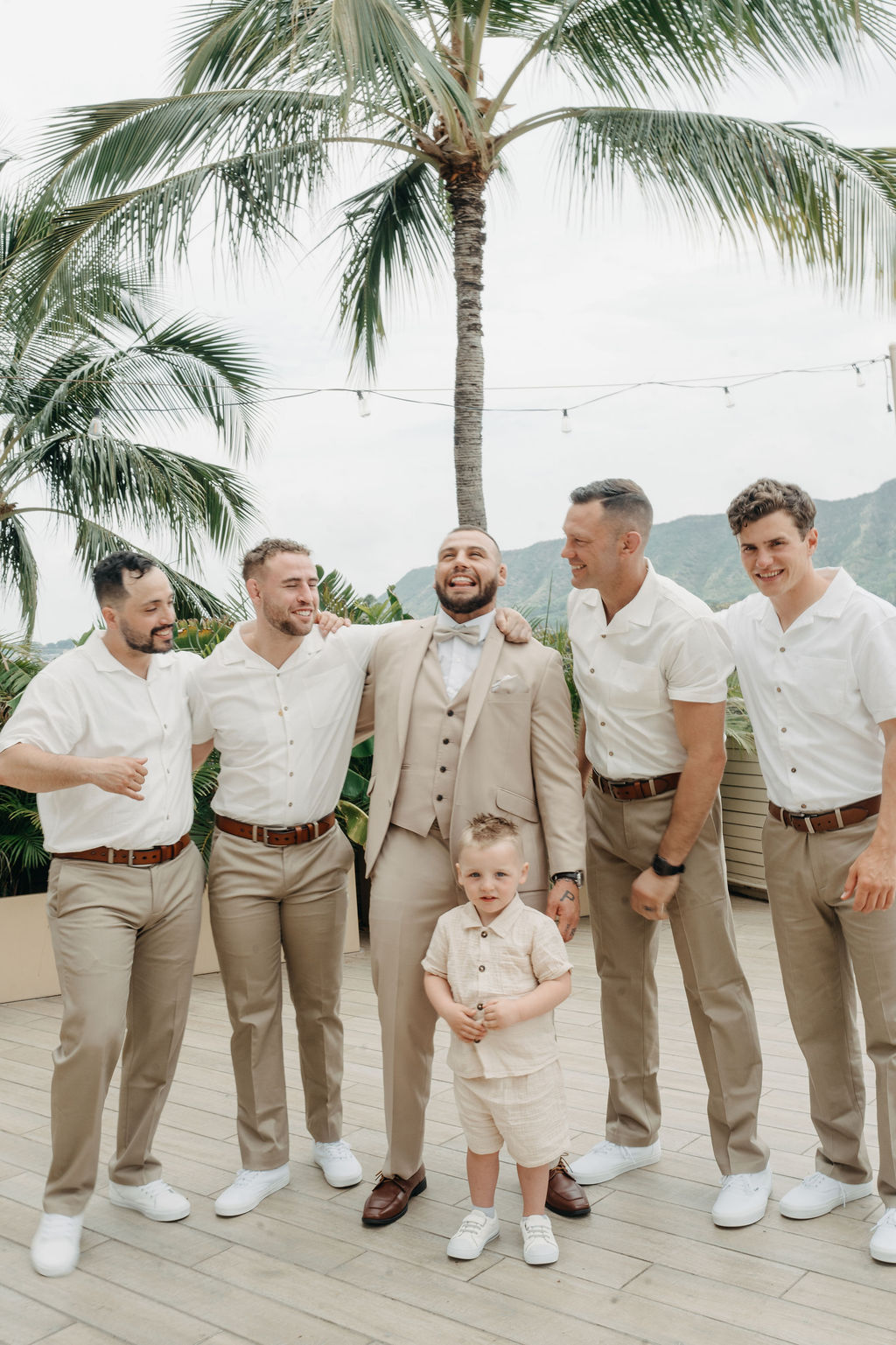 A bride and groom stand close together outdoors, surrounded by their wedding party in yellow and orange attire, with palm trees and mountains in the background for a wedding at Queen Kapiolani Hotel