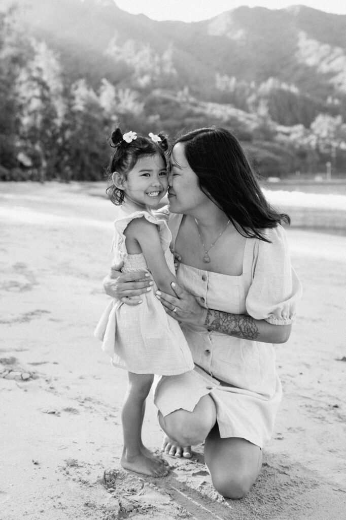 A woman kneels on a sandy beach, hugging and smiling at a young girl. Trees and mountains are visible in the background. The image is in black and white.