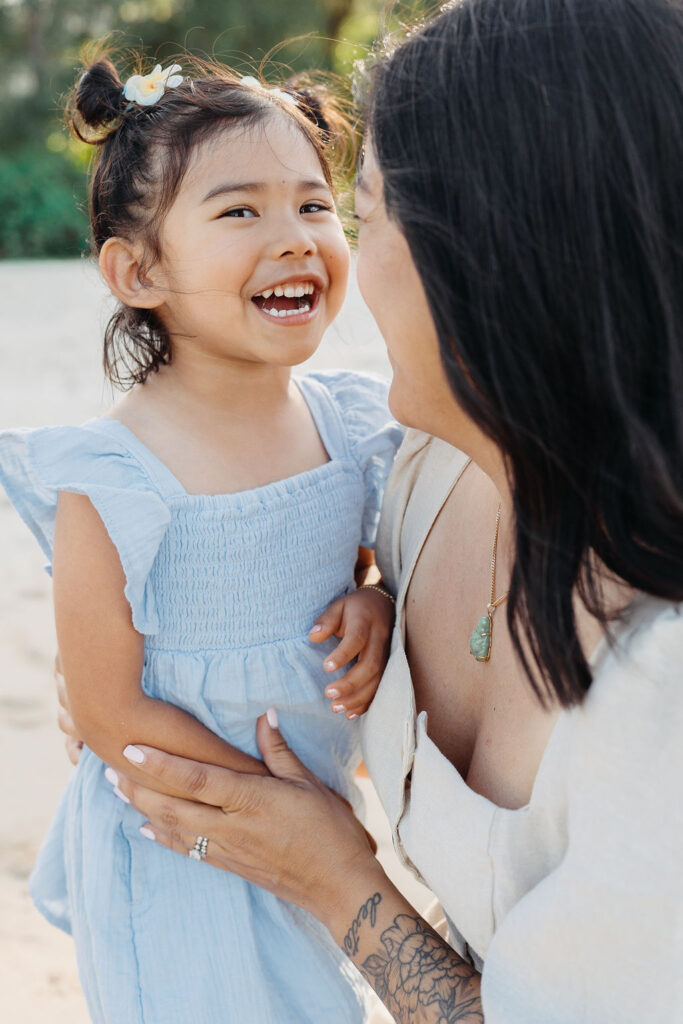 A woman kneels on a sandy beach, hugging and smiling at a young girl. Trees and mountains are visible in the background. 