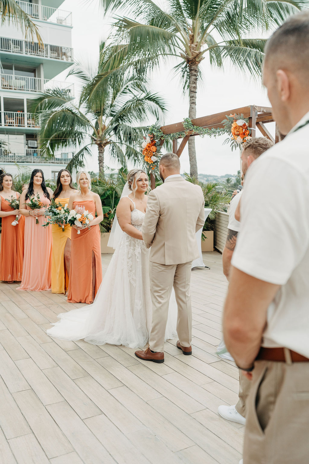 A bride and groom stand facing each other during an outdoor wedding ceremony, with bridesmaids in orange dresses and palm trees in the background.