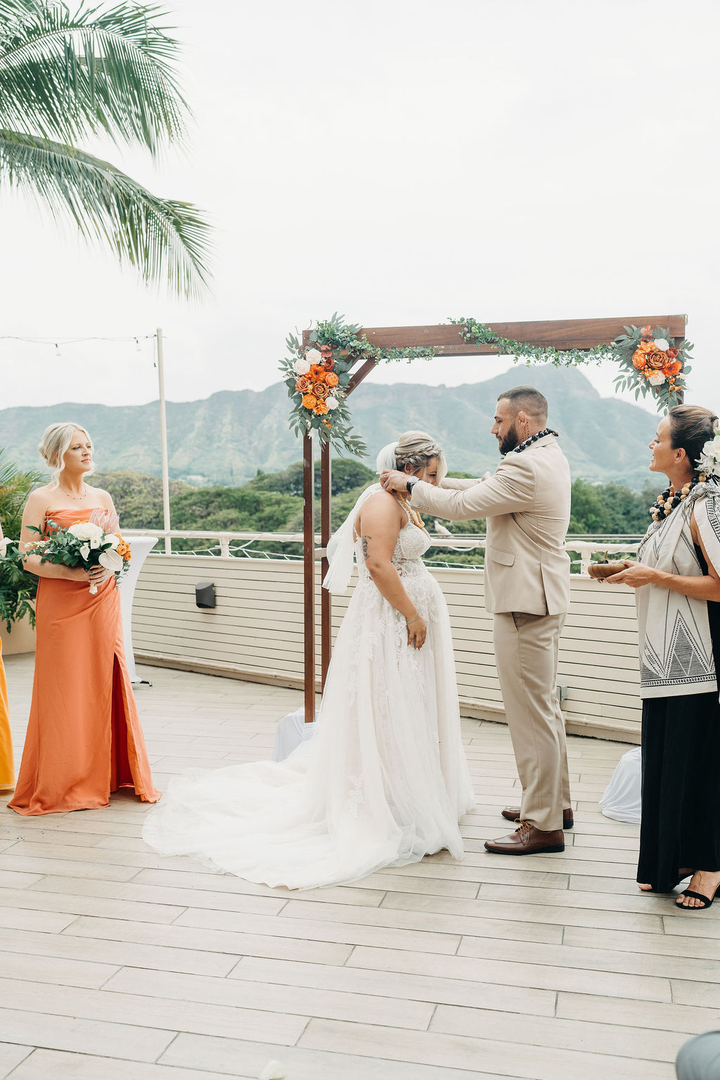 A bride and groom stand facing each other during an outdoor wedding ceremony, with bridesmaids in orange dresses and palm trees in the background.