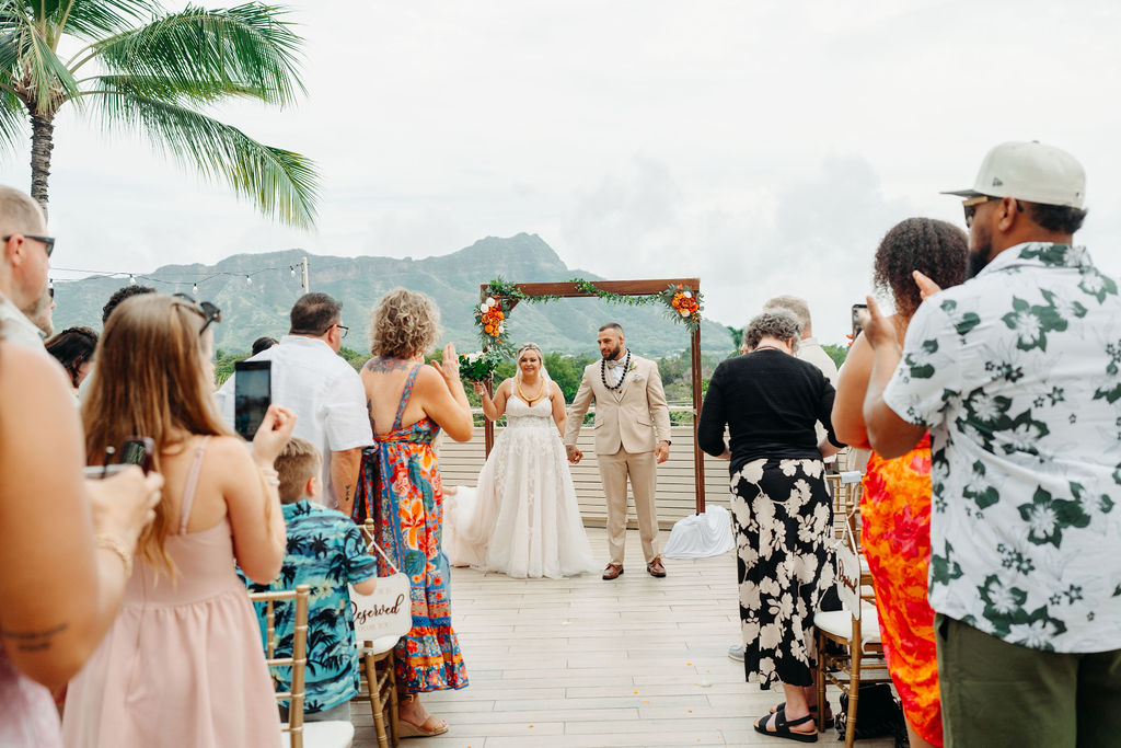 A bride and groom stand facing each other during an outdoor wedding ceremony, with bridesmaids in orange dresses and palm trees in the background.