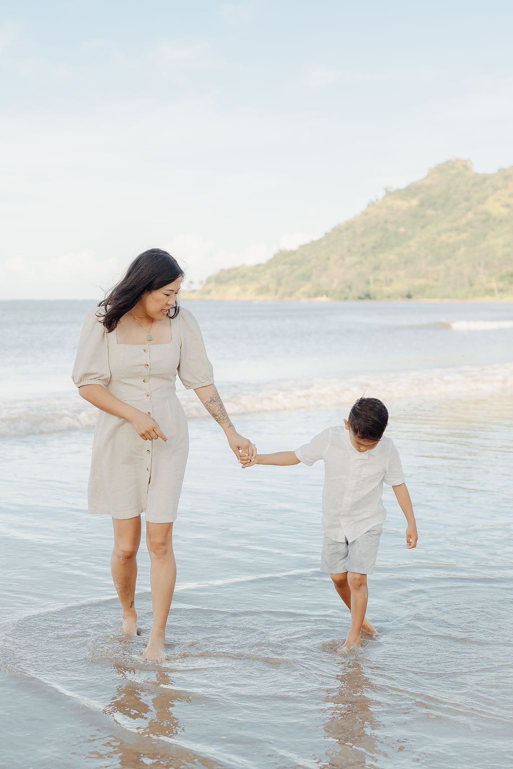 A woman and a young boy walk barefoot along the shoreline, holding hands, with gentle waves and a green hill in the background.
