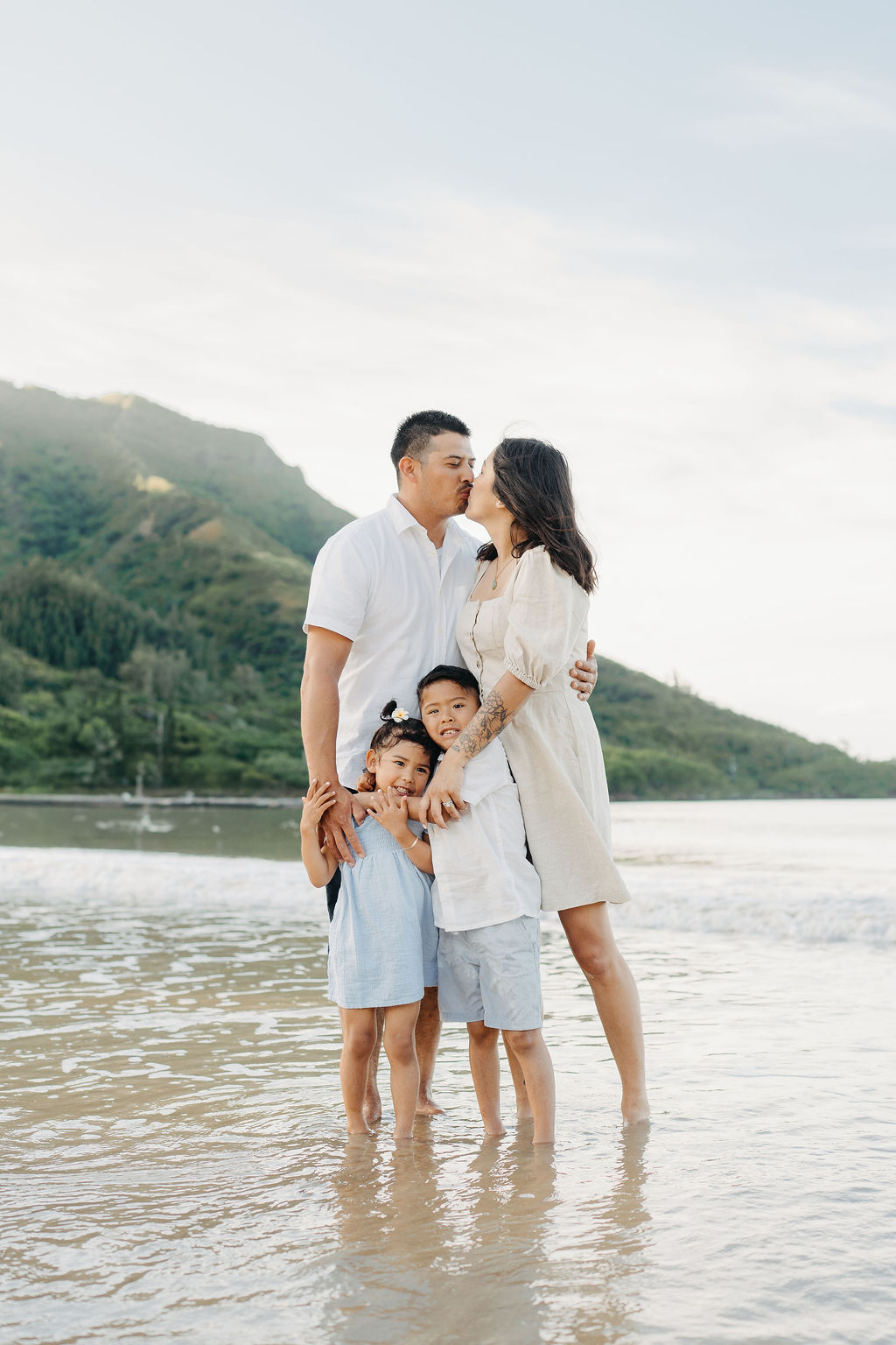 A family of four stands together on a beach with mountains in the background, dressed in light-colored clothing, smiling at the camera.