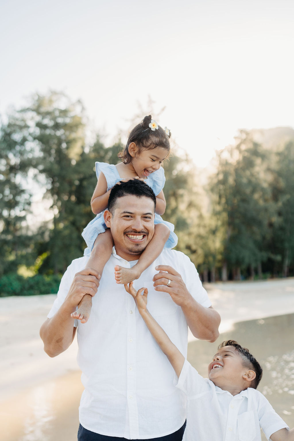 A man smiles while carrying a young girl on his shoulders, with a boy standing beside him outdoors near trees and water.