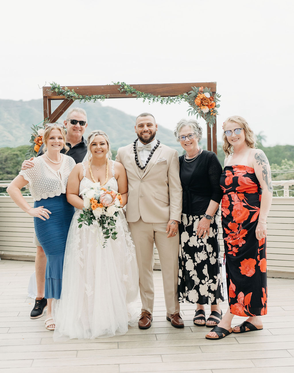 A bride and groom stand with their family in front of a wooden wedding arch decorated with greenery and flowers, outdoors with a mountain view.