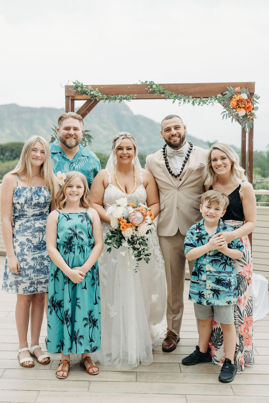 A bride and groom stand with their family in front of a wooden wedding arch decorated with greenery and flowers, outdoors with a mountain view.