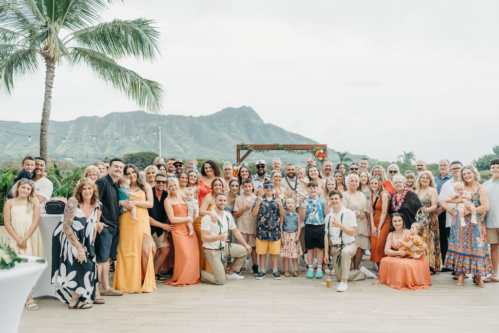 A bride and groom stand with their family in front of a wooden wedding arch decorated with greenery and flowers, outdoors with a mountain view.