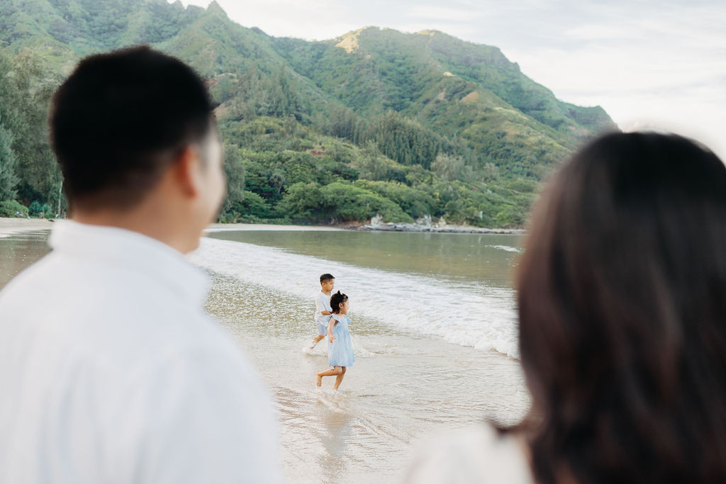 A child in a white shirt and shorts stands in shallow water at the beach, smiling as water splashes around, with green mountains in the background.