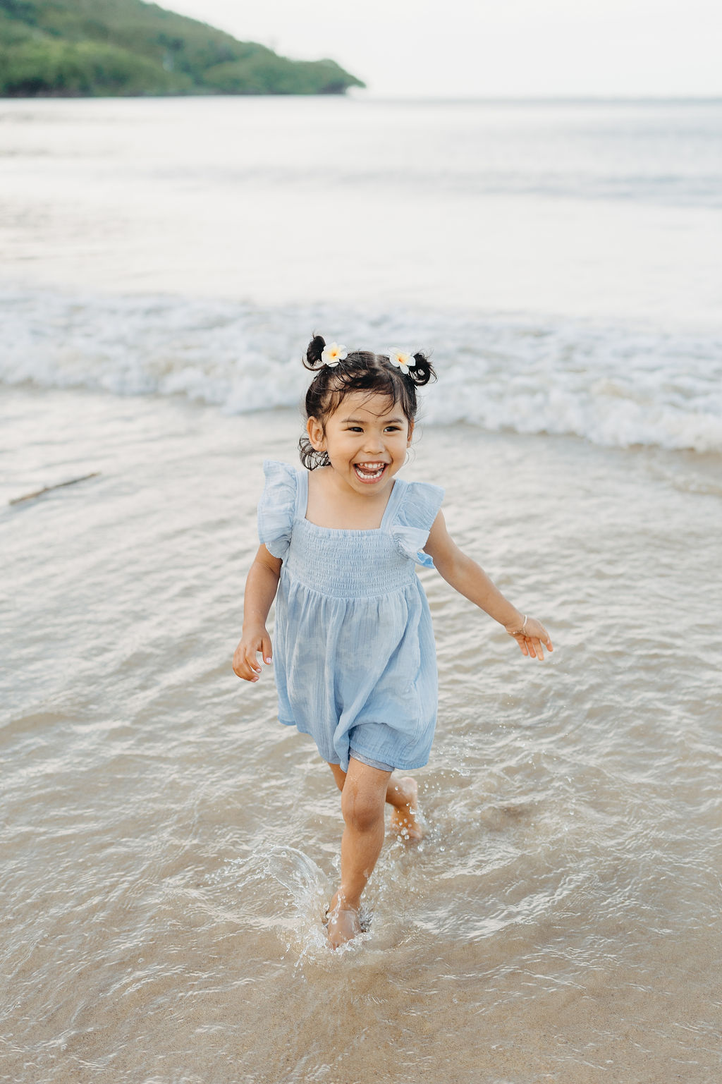 A young child in a light blue dress runs through shallow water at the beach, smiling, with gentle waves in the background.