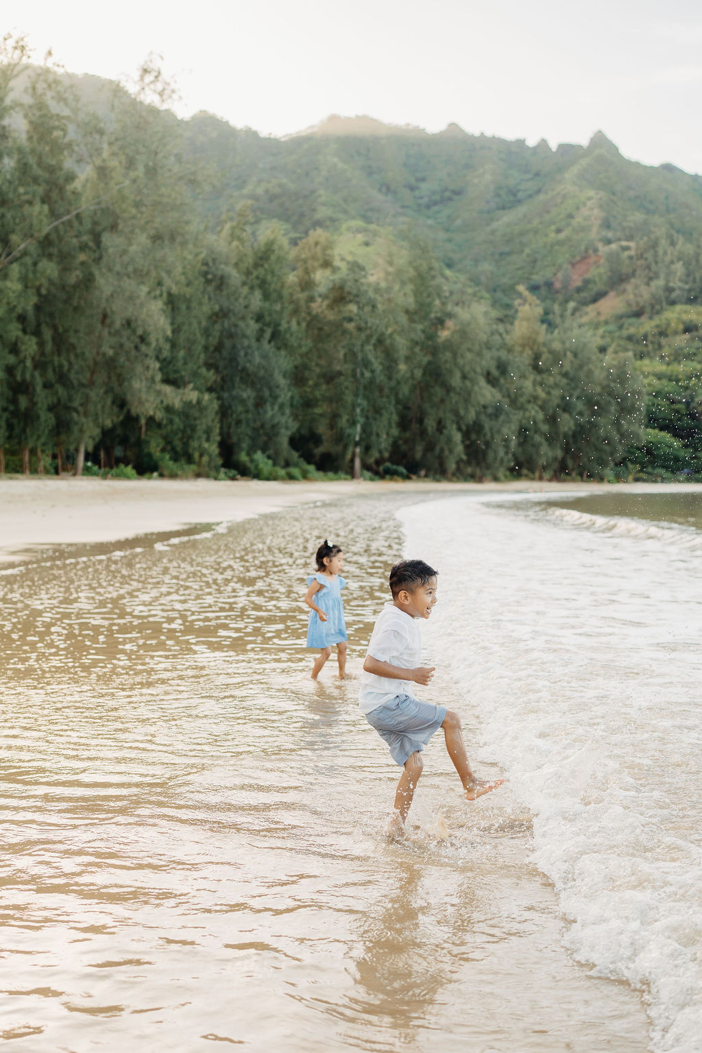Two children play in shallow water at the edge of a beach, with trees and green hills in the background.