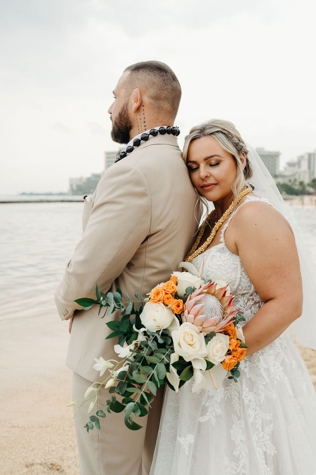 Bride and groom stand on a sandy beach with palm trees and city buildings in the background, holding a bouquet of flowers and wearing formal wedding attire or a wedding at Queen Kapiolani Hotel 