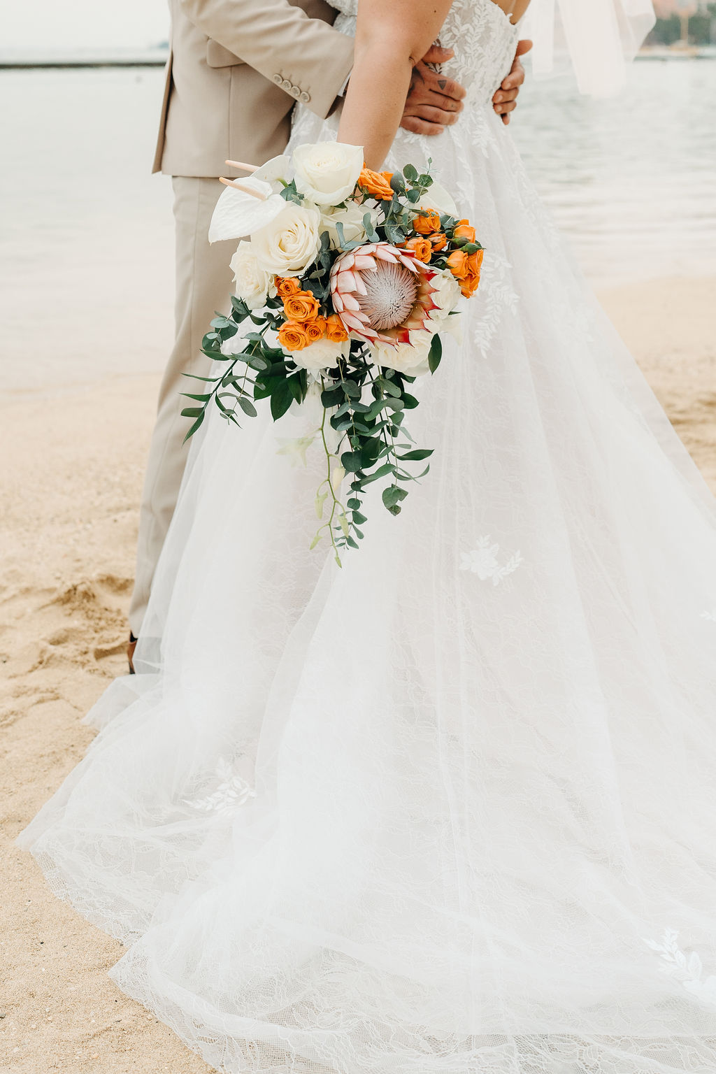 Bride and groom stand on a sandy beach with palm trees and city buildings in the background, holding a bouquet of flowers and wearing formal wedding attire or a wedding at Queen Kapiolani Hotel