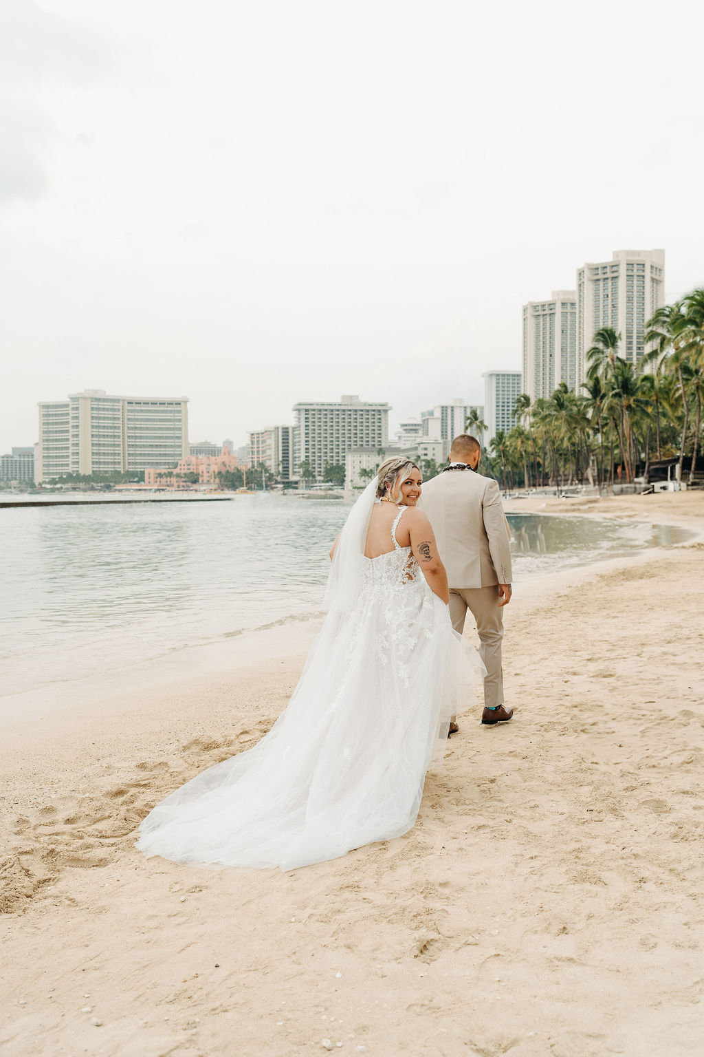 Bride and groom stand on a sandy beach with palm trees and city buildings in the background, holding a bouquet of flowers and wearing formal wedding attire or a wedding at Queen Kapiolani Hotel 