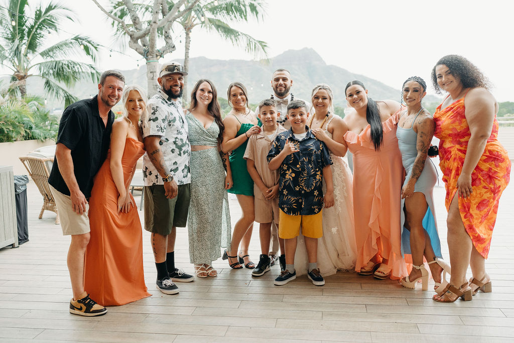 A bride and groom stand with their family in front of a wooden wedding arch decorated with greenery and flowers, outdoors with a mountain view.
