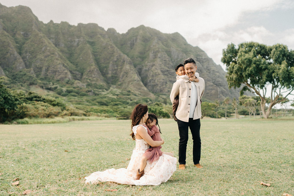 A family of four poses together on a grassy field with green mountains in the background for their mini family session