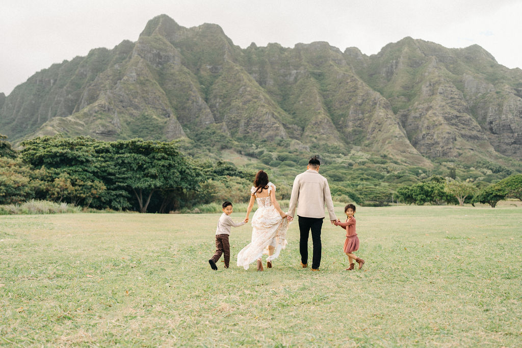 A family of four poses together on a grassy field with green mountains in the background for their mini family session