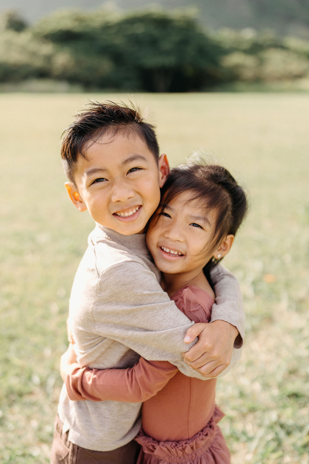 Two young children, a boy and a girl, stand outside on grass and smile while hugging each other. Trees and greenery are blurred in the background.